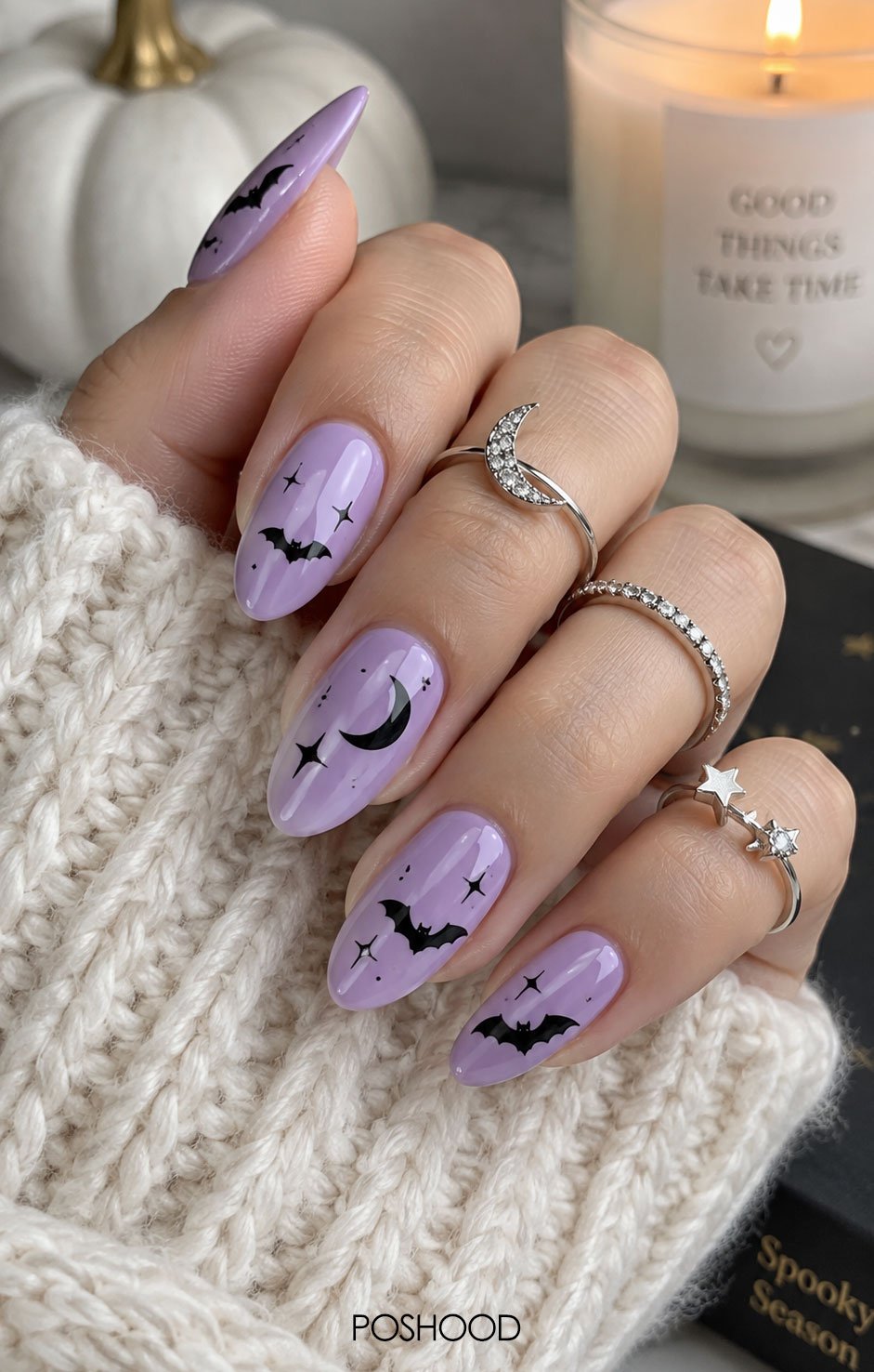 Glossy lavender almond-shaped Halloween nails with black bats, stars, and moon accents, styled with cream knit sleeves and silver rings.