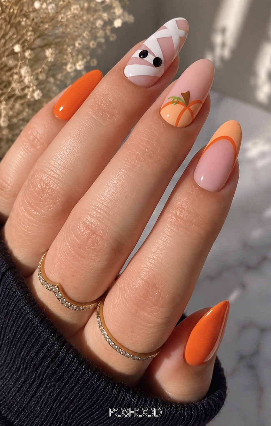Short almond-shaped nude Halloween nails decorated with white mummy wraps and playful googly eyes, styled with beige knit sleeves.