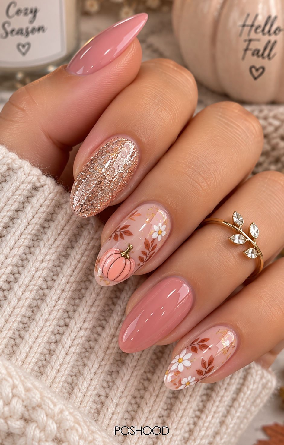 Glossy pink almond-shaped nails decorated with tiny blush pumpkins, delicate autumn leaves, gold foil, and subtle glitter, styled with cozy cream knitwear.