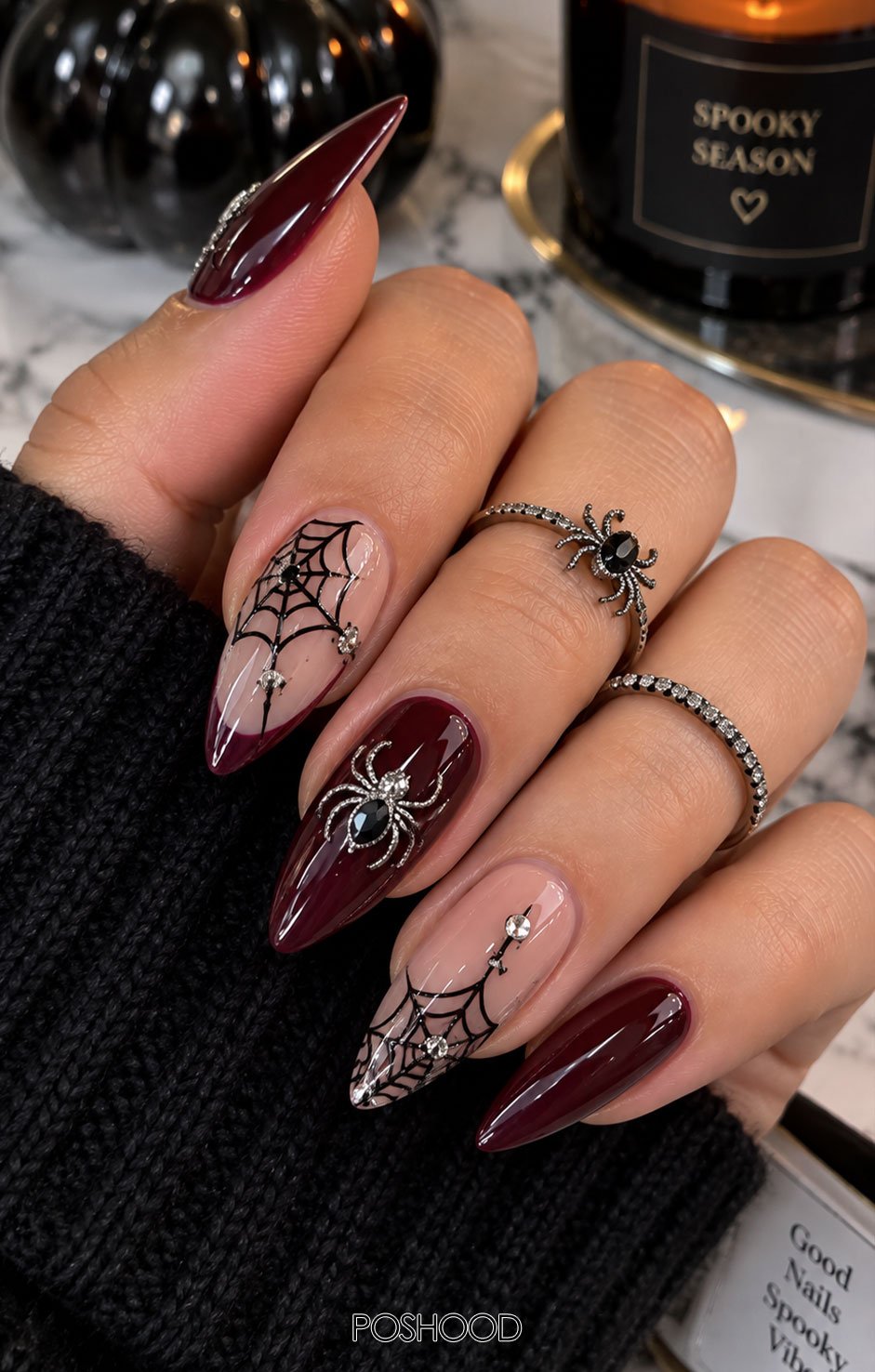 Long glossy almond-shaped Halloween nails with black spider webs, silver spider designs, crystal embellishments, and burgundy accents against a marble background.