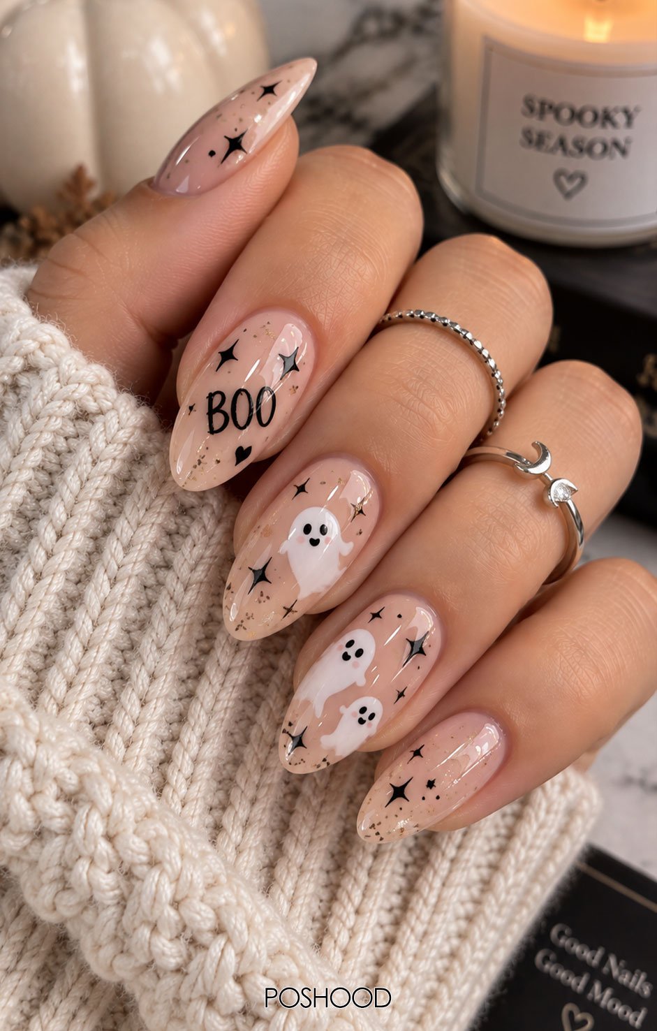 Glossy nude almond-shaped Halloween nails featuring BOO lettering, smiling white ghosts, black stars, and subtle glitter accents, styled with chunky knit sleeves and silver rings.