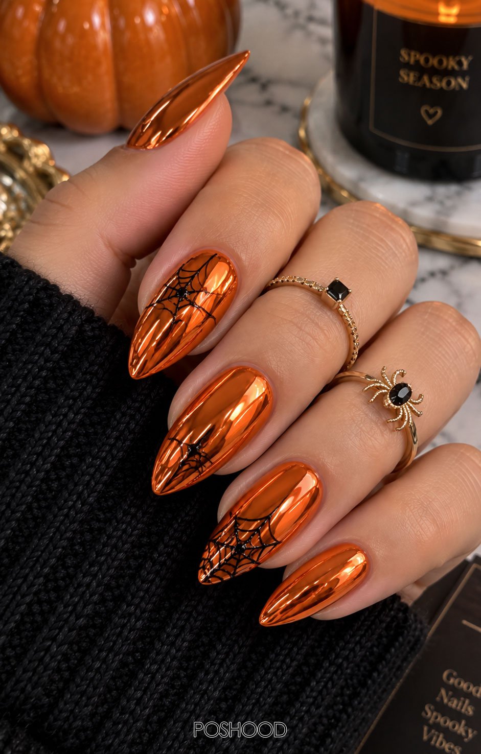 Glossy chrome orange almond-shaped Halloween nails featuring delicate black spider webs and metallic reflections, styled with elegant gold rings against a marble background.