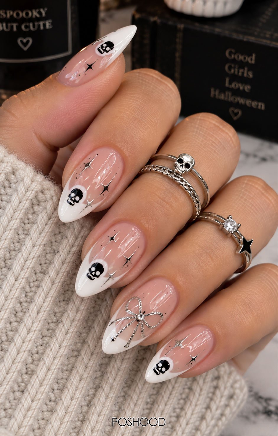 Glossy nude almond-shaped Halloween nails with white French tips, tiny black skulls, miniature stars, and silver glitter accents, styled with cream knit sleeves and silver rings.