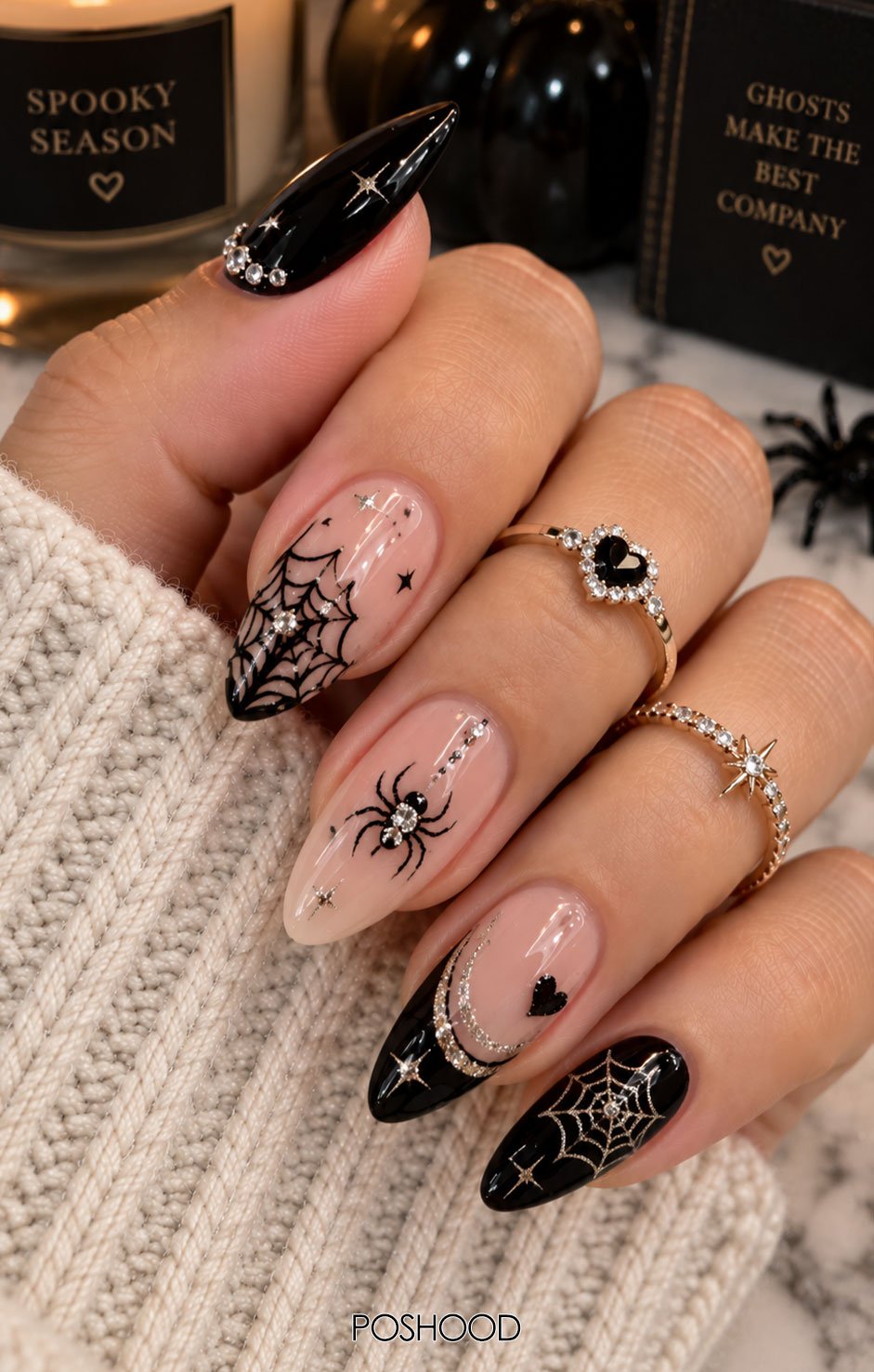 Glossy nude and black almond-shaped Halloween nails featuring delicate spider webs, a crystal spider, silver glitter accents, and elegant rings against a cozy neutral background.