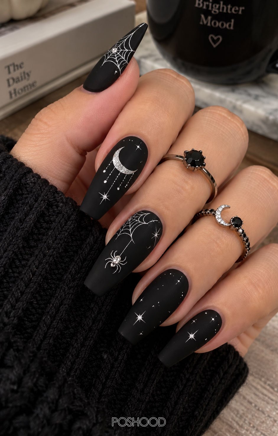 Long matte black coffin-shaped Halloween nails decorated with delicate spider webs, crescent moons, and minimalist white stars, styled with a black knit sleeve and silver rings against a natural wooden background.
