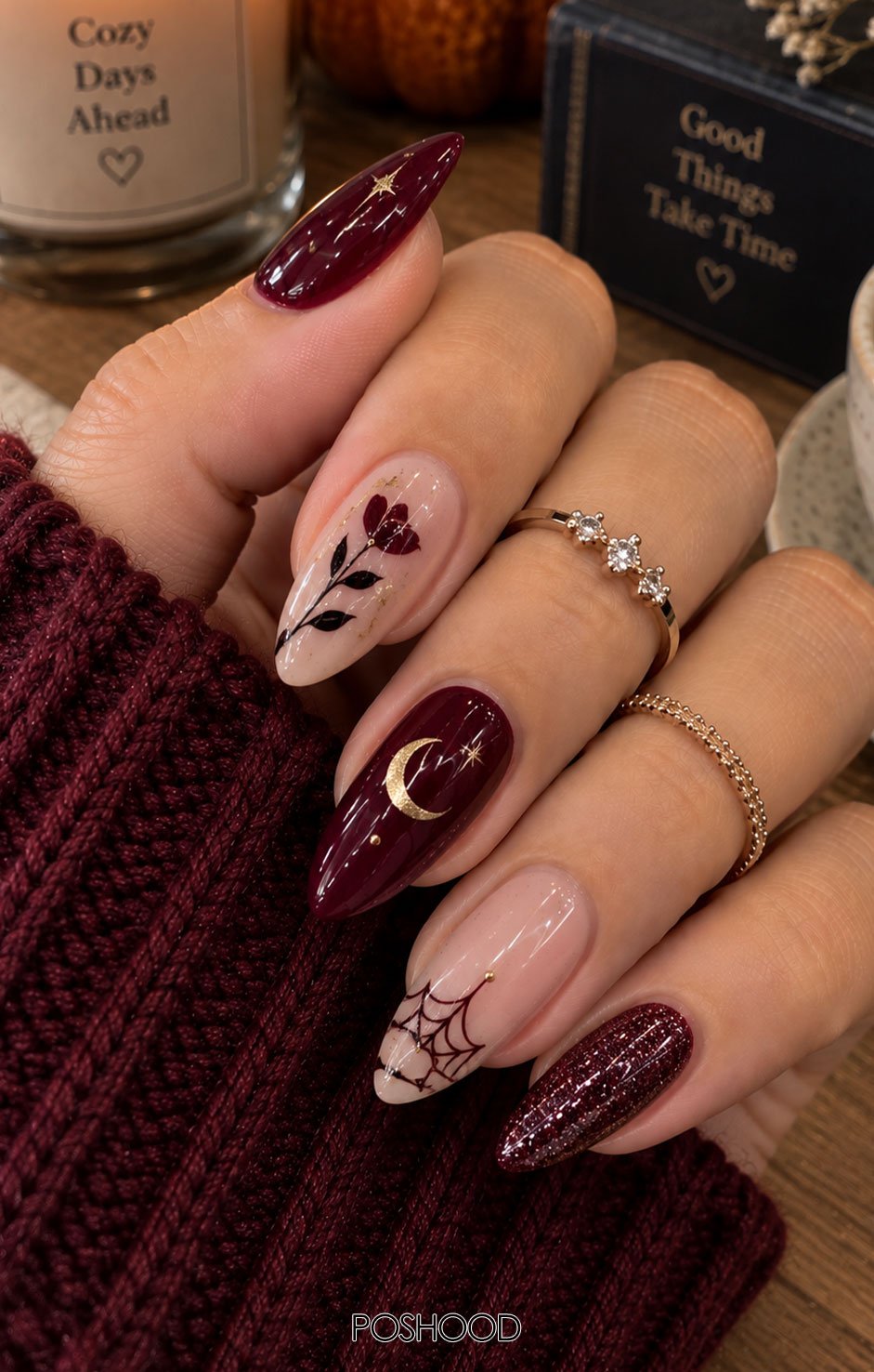 Glossy burgundy almond-shaped Halloween nails featuring floral designs, black leaves, gold accents, crescent moons, and delicate spider webs, styled with cozy knitwear against a warm autumn background.