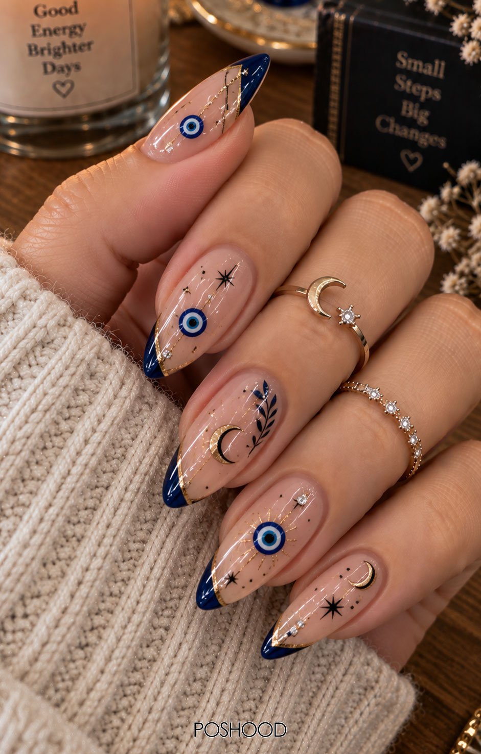 Glossy nude almond-shaped nails featuring blue evil-eye designs, navy French tips, crescent moons, black stars, gold accents, and tiny crystals, styled with cozy cream knitwear and gold rings.