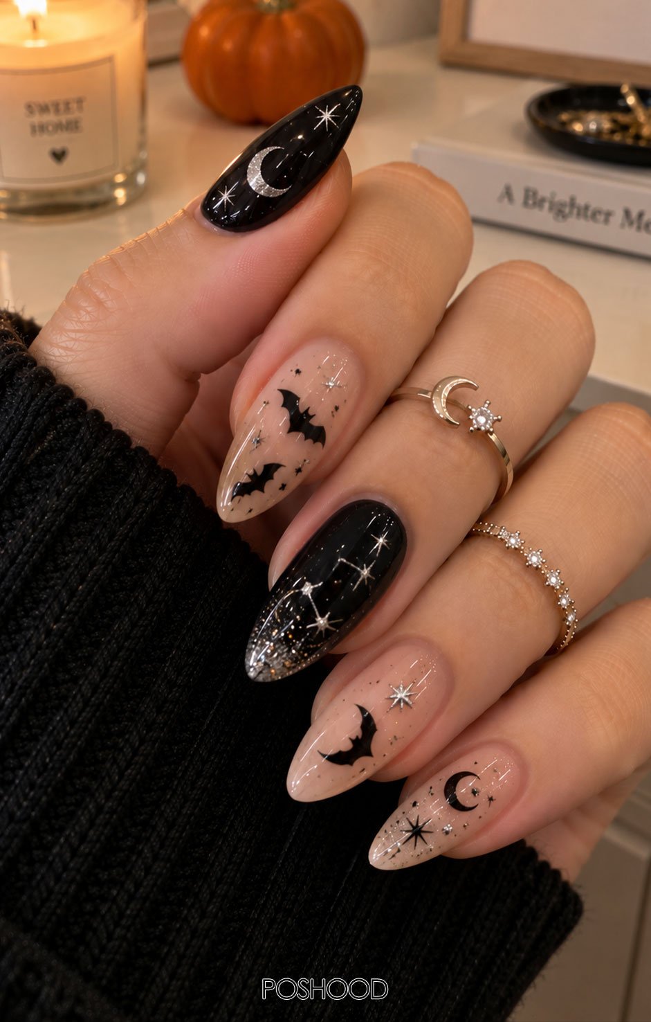 Glossy black and nude almond-shaped Halloween nails featuring tiny bats, crescent moons, silver stars, sparkling constellations, and delicate glitter, styled with cozy black knitwear and silver rings in a natural room setting.